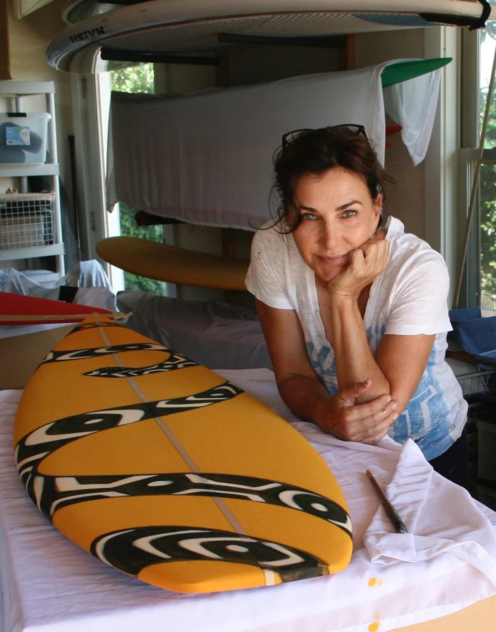 Kim McDonald leans against a workbench inside a surfboard workshop, with a yellow surfboard featuring black and white abstract patterns in the foreground.