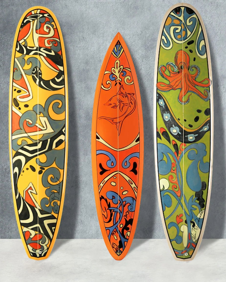 Three decorative surfboards with colorful abstract and tribal-style patterns leaning against a gray wall next to a teal sofa with patterned pillows.