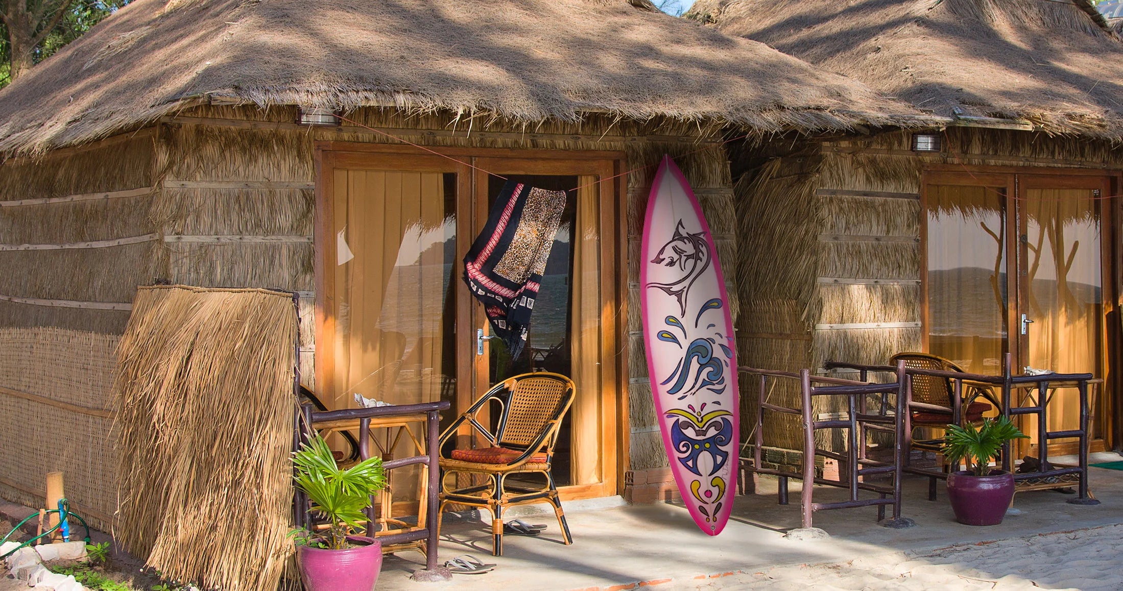 A rustic thatched-roof hut with wooden doors and outdoor seating, featuring a pink and white surfboard leaning against the wall.