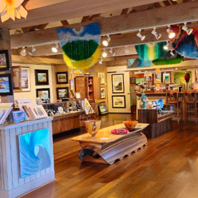 Hana Coast Gallery - A rustic art and craft shop interior with wooden floors, exposed beams, hanging colorful textile wall art, framed pictures on the walls, and display tables with small handmade items.