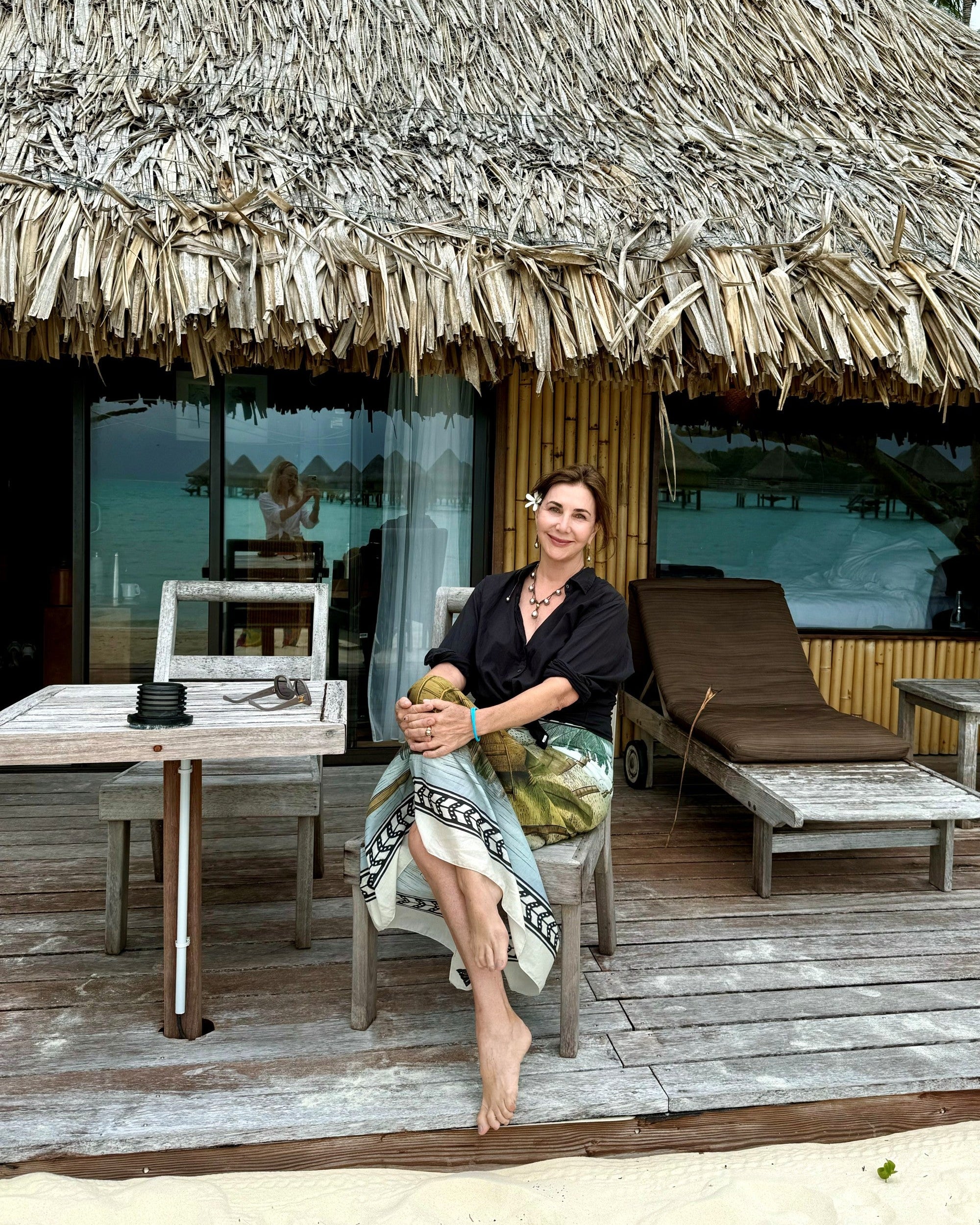 Kim sits barefoot on a wooden deck in front of a thatched-roof bungalow, holding a coconut. She wears a black short-sleeve top and a patterned skirt.