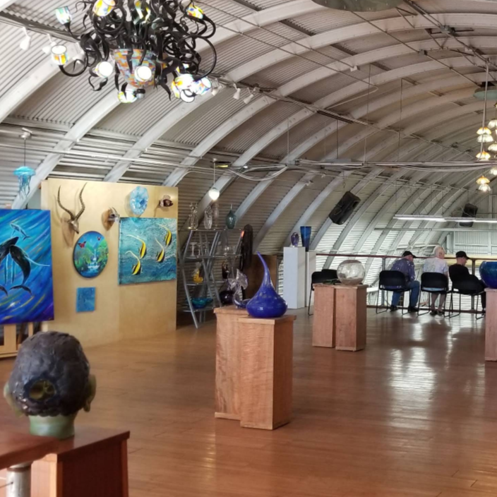 Makai Glass Maui - An indoor art gallery and glass blowing studio, with a curved metal roof, wood floors, hanging chandeliers, and multiple displayed artworks on pedestals and walls. 
